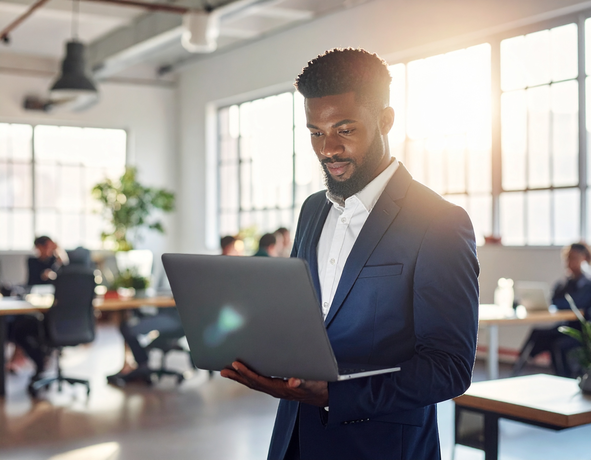 Homem elegante em terno azul trabalha em laptop em escritório moderno e iluminado pelo sol, transmitindo foco e profissionalismo. Ambiente inspirador e colaborativo com colegas ao fundo.
